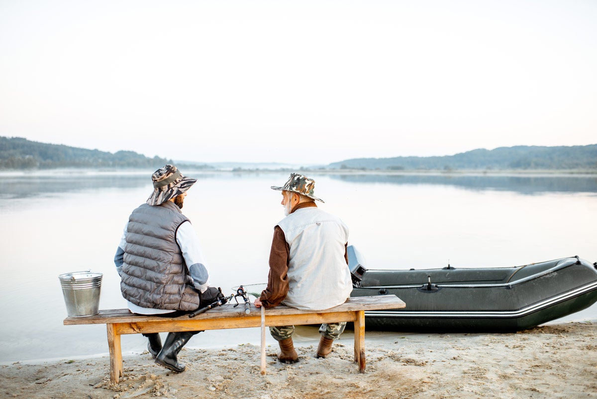 Grandfather with son sitting together on the bench while fishing on the lake early in the morning, back view