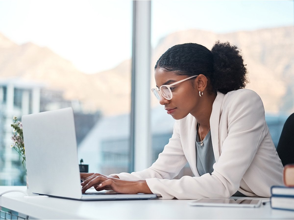 Woman with glasses working on laptop with mountains out the window in the back