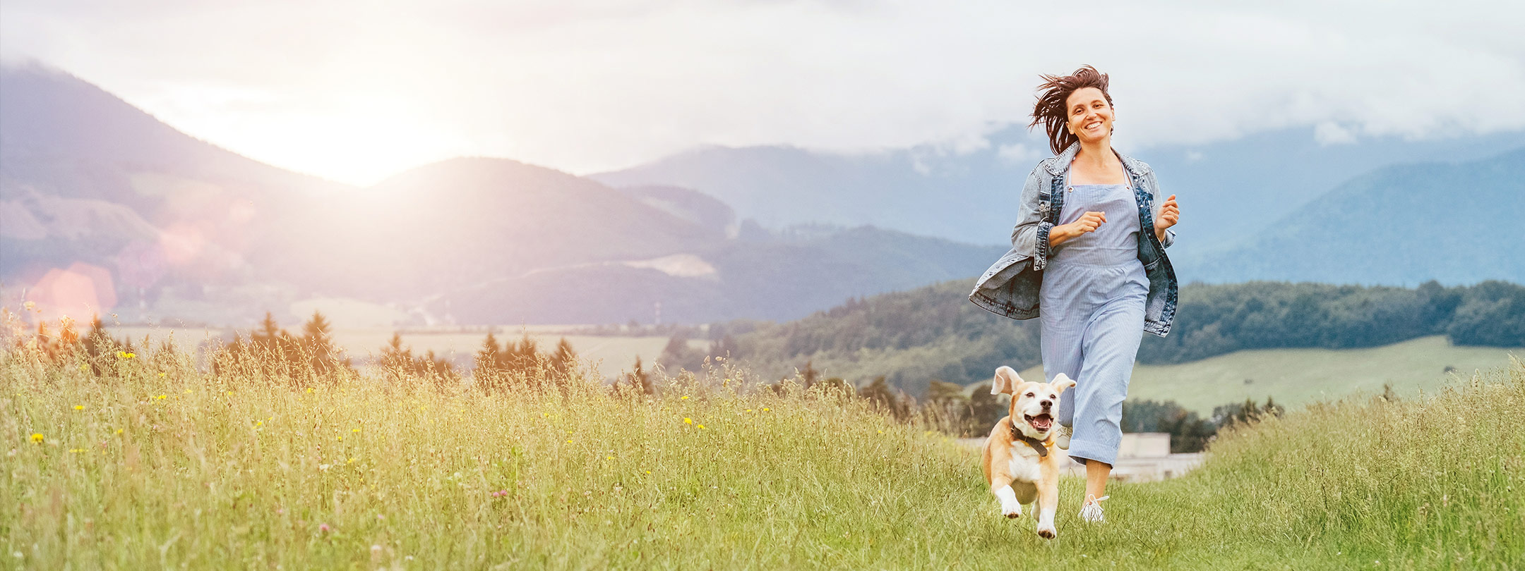 A young woman and her dog run through a field in a mountain meadow
