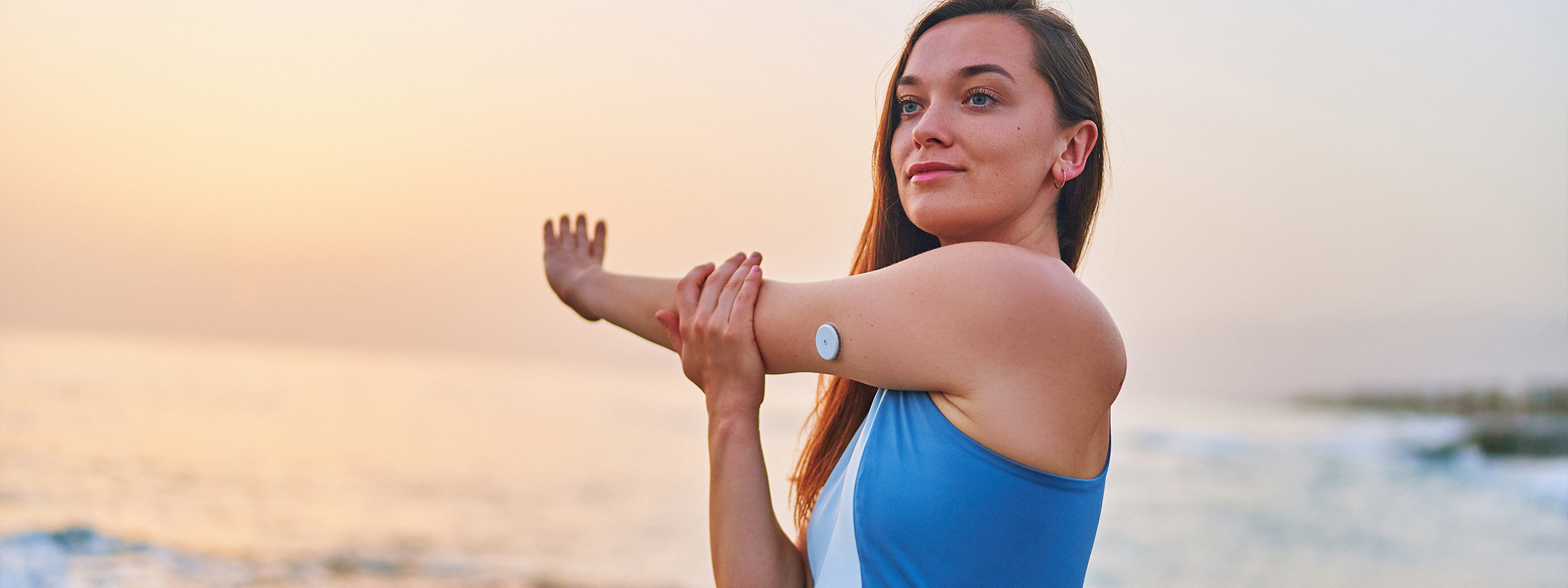 A woman stretches on a beach wearing a swimsuit and a blood sugar monitor