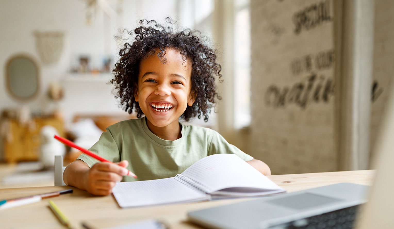 Child smiling at a desk with paper and colored pencils, looking at camera