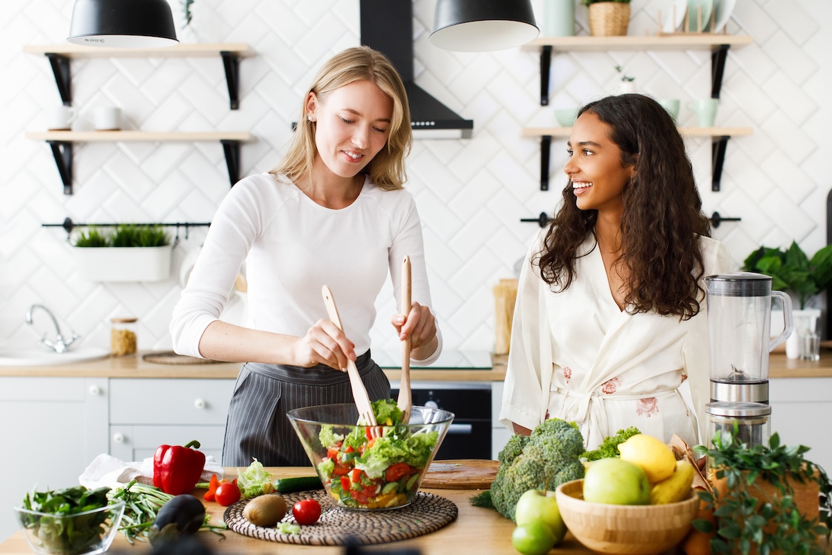 Attractive caucasian girl is cooking healthy salad and beautiful mulatto girl is looking  on her dressed in silky nightgown on modern designed kitchen