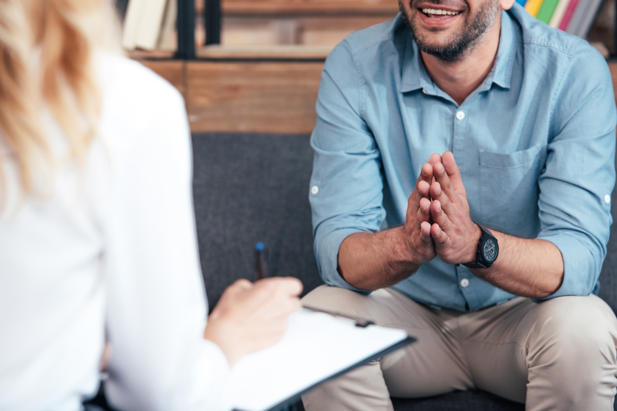 cropped image of female counselor writing in clipboard and smiling male holding hand palms together during therapy session in office 