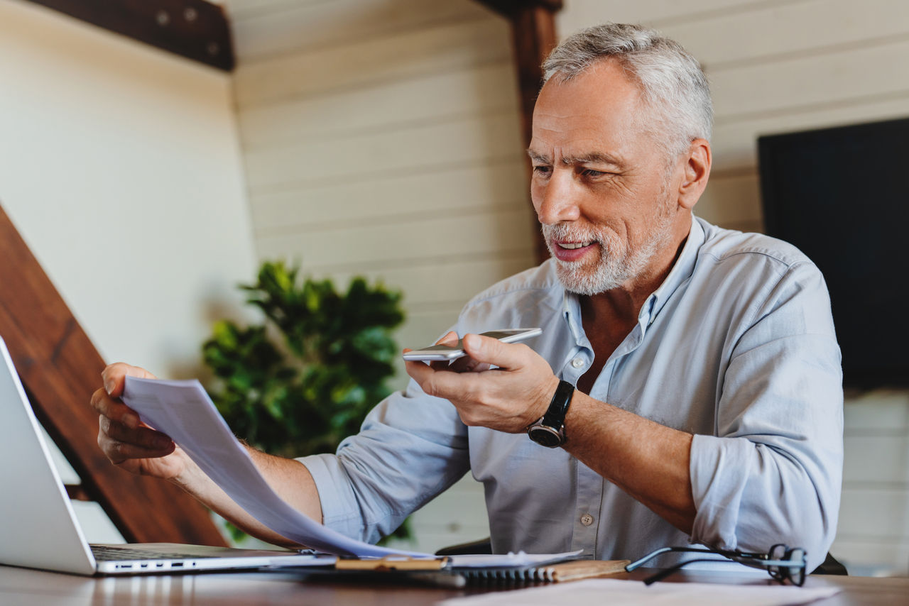 Senior man working at home while discussing using speakerphone
